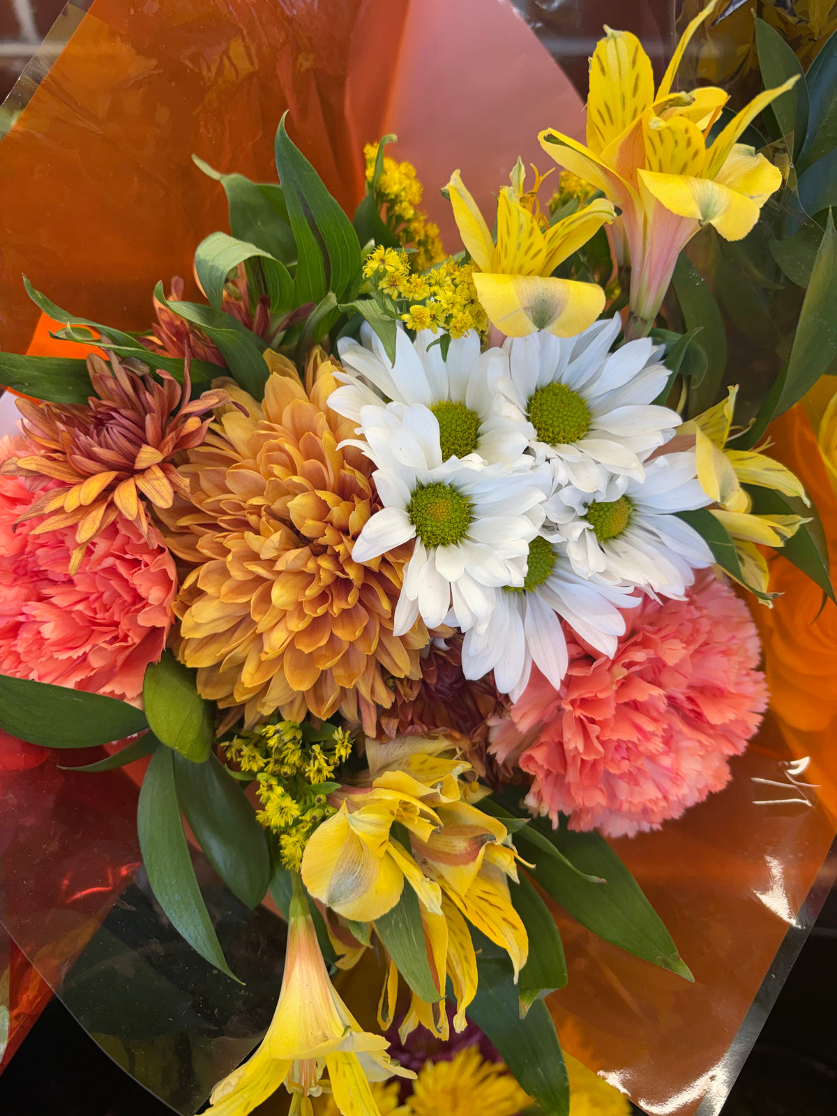 Mixed bouquet of white daisy-style flowers, peach carnations, golden chrysanthemums, and yellow alstroemeria in orange wrapping